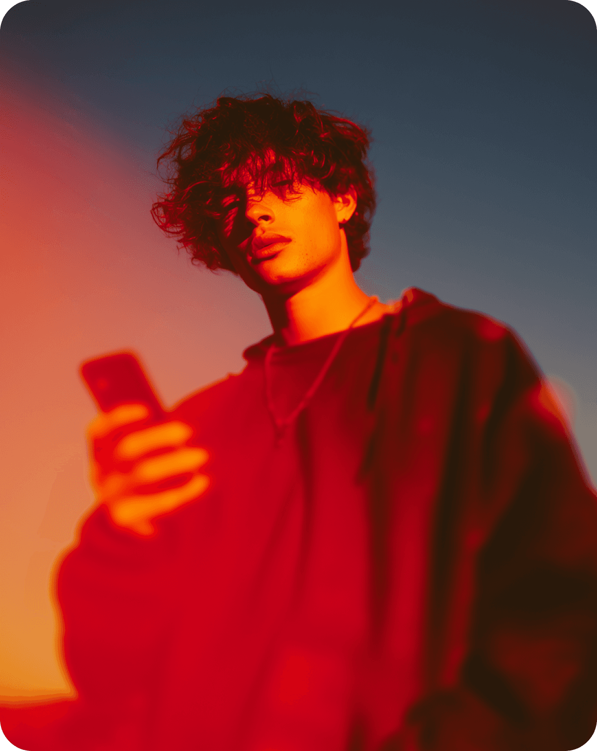 Jeune personne aux cheveux bouclés tenant un téléphone, éclairée de rouge et de bleu au crépuscule